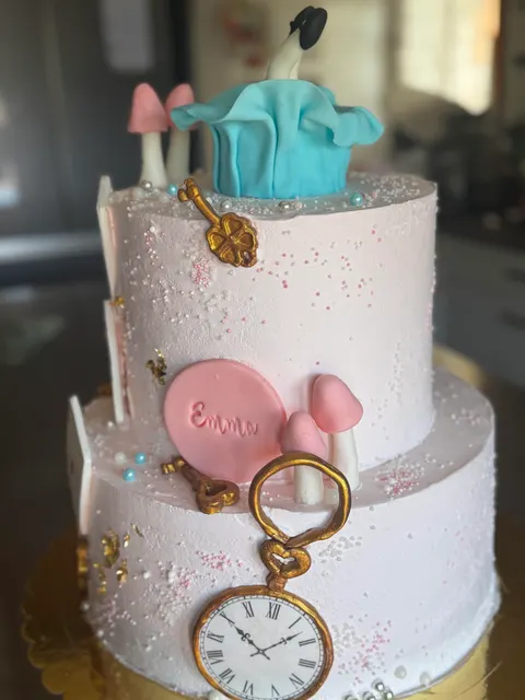 Torta scenografica a due piani ricoperta di crema rosa chiaro, decorata con chiavi dorate, un orologio da taschino, funghetti e Alice a testa ingiù sulla sommità.