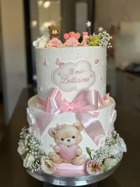 Torta a due piani bianca decorata con un grande fiocco rosa, un orsetto modellato in pasta di zucchero, cuori e fiori freschi, con la scritta Il mio battesimo.