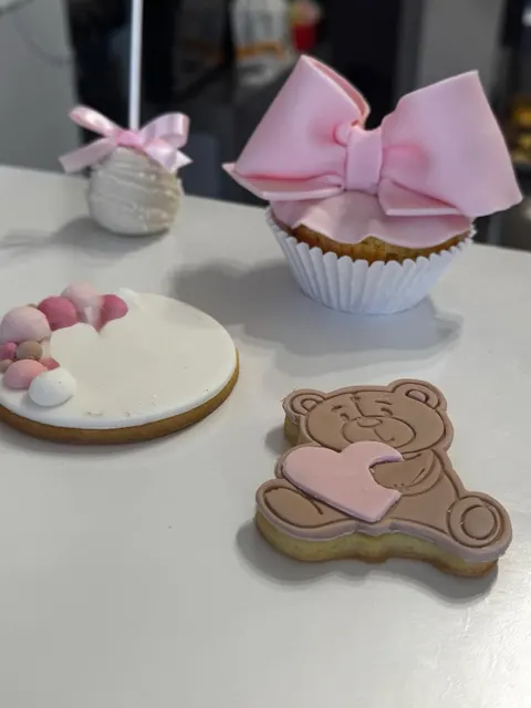 Set di dolcetti artigianali decorati con pasta di zucchero nei toni del rosa e bianco, tra cui un cupcake con grande fiocco, biscotti decorati con orsetto e cuori, e un cake pop.