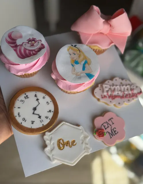 Set di cupcake decorati con Alice e lo Stregatto, biscotti a forma di orologio, fiocco in pasta di zucchero e targhette Eat Me su fondo bianco