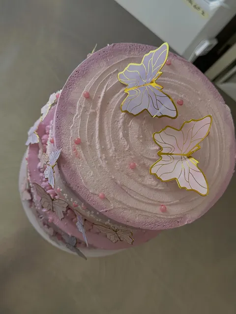 Vista dall'alto di una torta a più piani con finitura sfumata rosa e lilla, decorata con sagome di farfalle con bordi dorati e perline sferiche rosa sulla superficie e lungo i lati.
