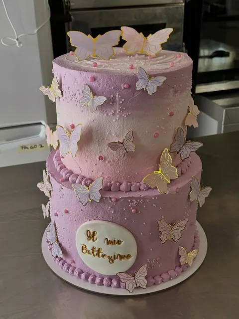 Torta a due piani rotonda con copertura rosa sfumata, decorata con numerose farfalle di carta o cialda bianche e rosa, palline di zucchero e una targa rotonda con scritta dorata Il mio Battesimo