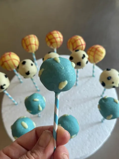 Una mano tiene un cake pop azzurro con macchie chiare su un bastoncino a righe bianche e azzurre; sullo sfondo altri cake pops con fantasie gialle a quadretti e bianche macchiate di nero.