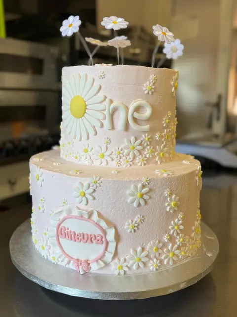 Torta a due piani di colore rosa chiaro, ricoperta di margherite bianche con centro giallo. Sul piano superiore c'è la scritta bianca one e sul piano inferiore un medaglione con il nome Ginevra in rilievo