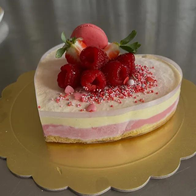Torta a forma di cuore poggiata su un vassoio dorato, con strati di colore giallo chiaro e rosa a vista sui lati. Sulla superficie superiore sono disposti lamponi, fragole con foglioline, un pasticcino tondo rosa e piccoli zuccherini colorati a forma di cuore e palline.