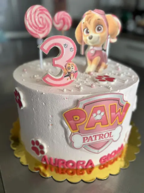 Torta rotonda con copertura rosa chiaro, decorata con il logo Paw Patrol sul davanti, il numero tre rosa, sagome di cagnolini e lecca-lecca bianchi e rossi sulla sommità
